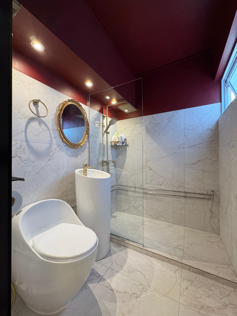 HDB bathroom with white marble tiles, burgundy ceiling, gold ornate mirror and glass shower
