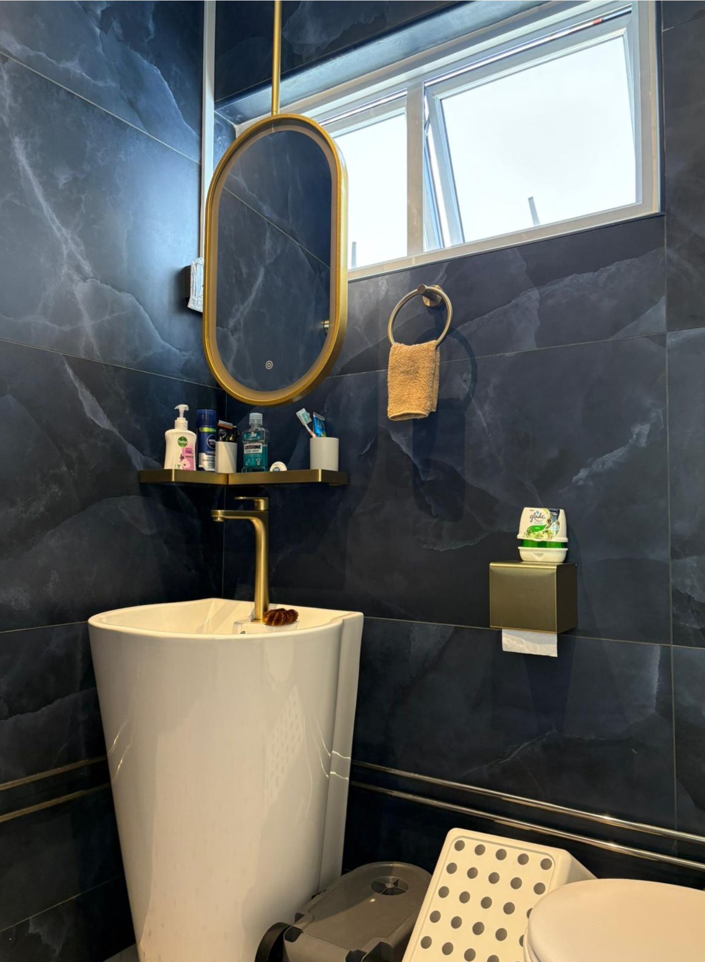 Bathroom with dark navy marble tiles, white pedestal basin, brass oval mirror and gold tap